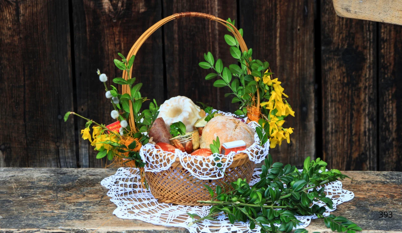 O tych zasadach pamiętaj przy świątecznym stole Wiklinowy koszyk ozdobiony białą koronką i zielonym bukszpanem, wypełniony tradycyjnymi pokarmami wielkanocnymi: jajkiem, chlebem, kiełbasą i ciastem, w otoczeniu świeżych gałązek i żółtych kwiatów, ustawiony na drewnianym stole.
