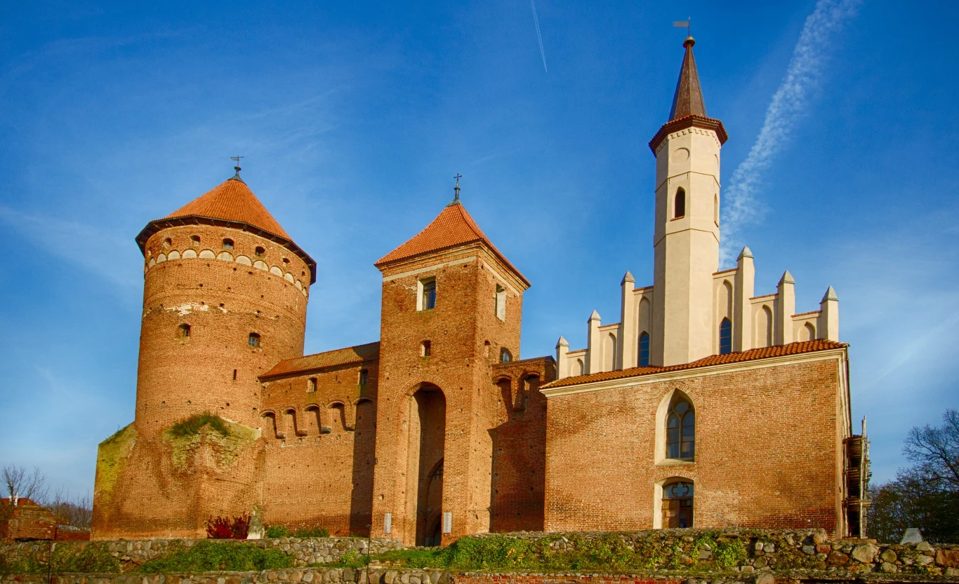 gotycki zamek z masywną okrągłą wieżą i wysoką smukłą wieżą kościelną, ceglany mur obronny, duże ostrołukowe okna, niebieskie niebo w tle