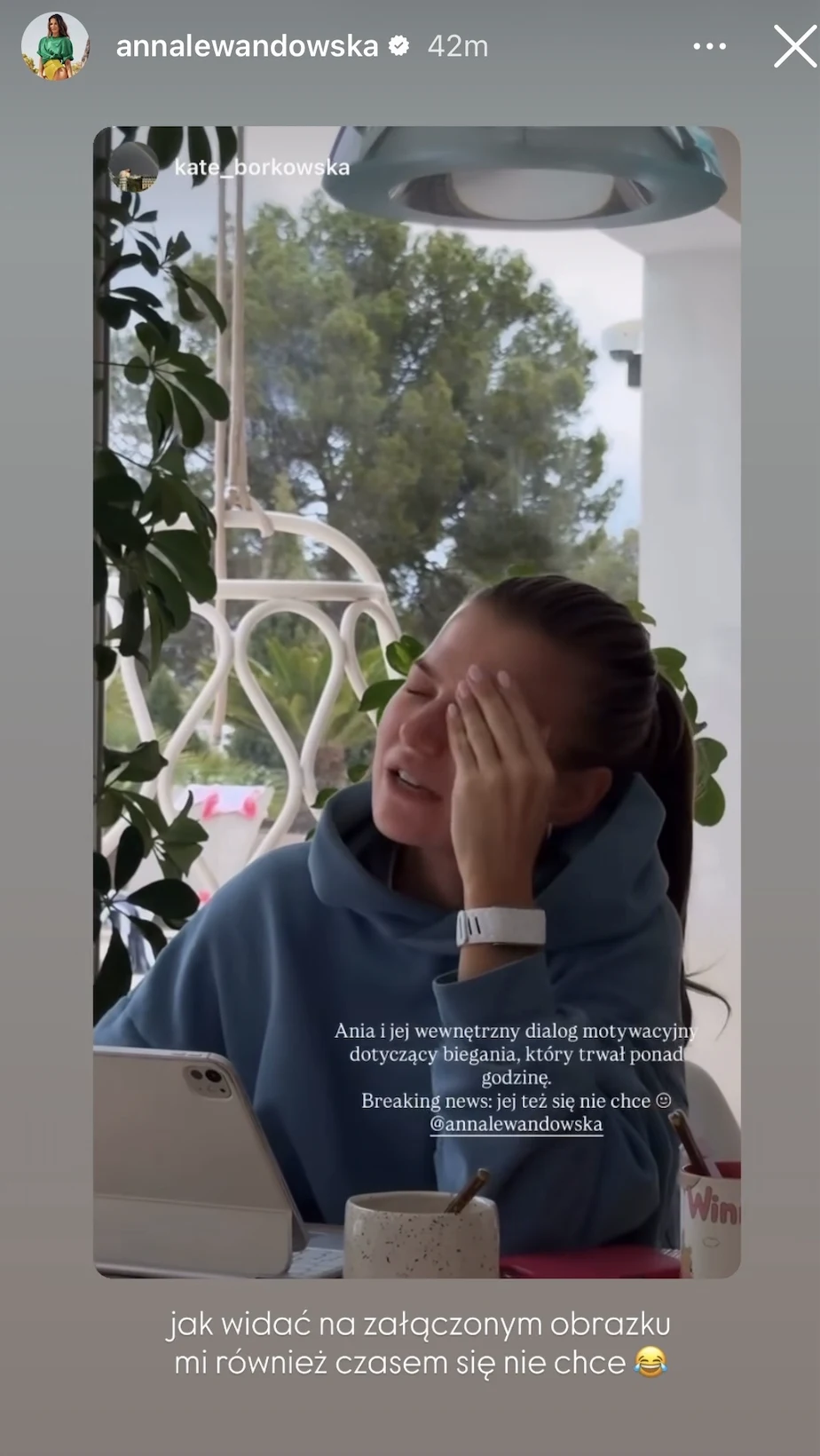 Kobieta siedzi przy stole na tarasie, wspiera głowę na dłoni i wygląda na zmęczoną lub zrezygnowaną, ubrana w niebieską bluzę, przed nią leży laptop, kubek oraz zeszyt, w tle widoczne są białe meble ogrodowe i zielone drzewa.