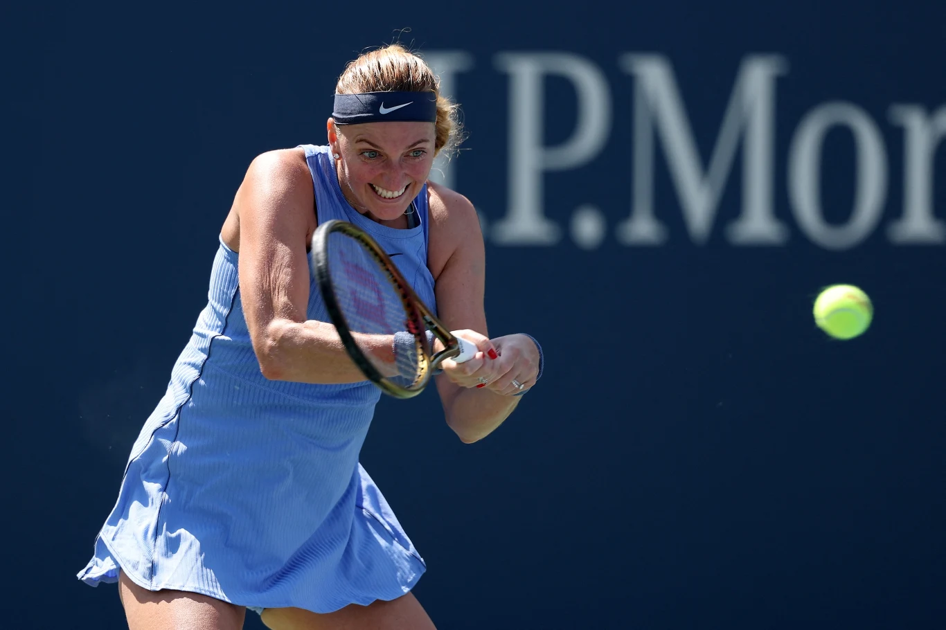 Tenisistka w niebieskiej sukience wykonuje mocny backhand dwuręczny, skupiona na piłce lecącej tuż przed nią, w tle widoczne fragmenty logotypu sponsora na ciemnoniebieskim tle.