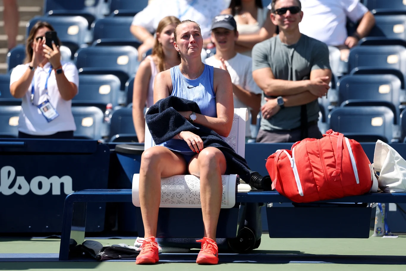 Zawodniczka tenisowa siedzi na ławce kortu, trzymając ręcznik na kolanach i wyglądając na wzruszoną, obok niej leży duża czerwona torba sportowa, w tle widzowie śledzą wydarzenie.