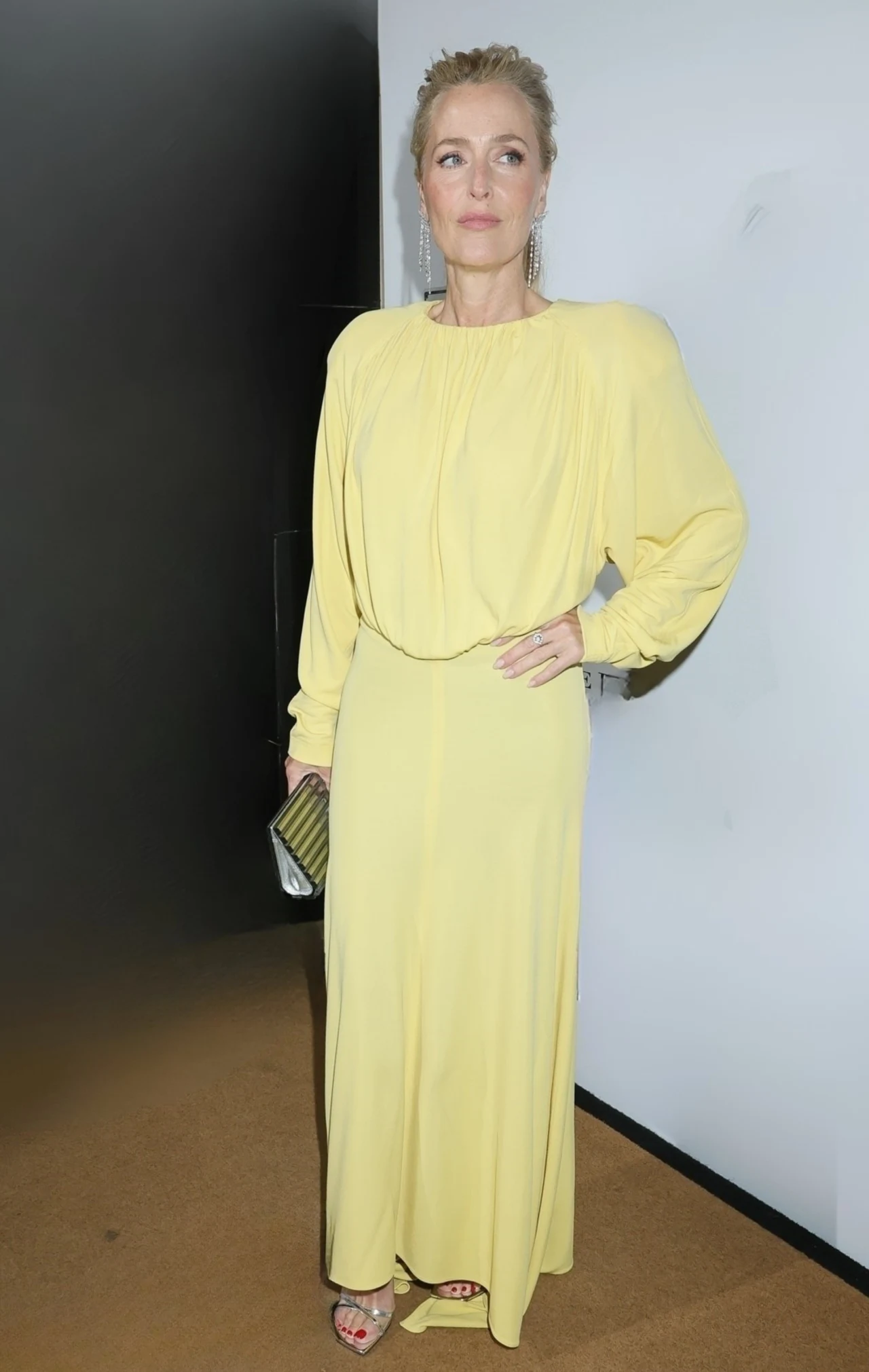 Gillian Anderson kobieta w długiej, jasnożółtej sukni z długimi rękawami, trzymająca metaliczną kopertówkę i mająca delikatny makijaż oraz upięte włosy, stojąca przy białej ścianie
