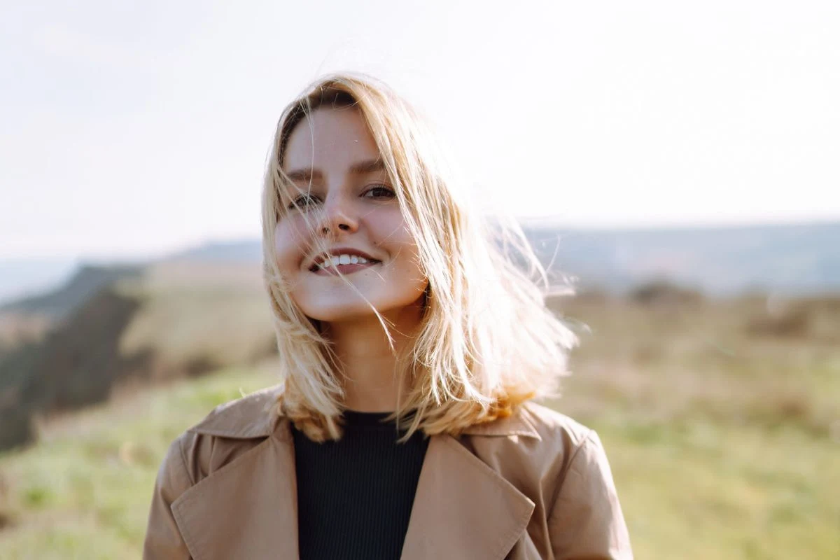 Młoda kobieta z blond włosami i szerokim uśmiechem, ubrana w jasnobrązowy płaszcz i czarną bluzkę, stoi na otwartej przestrzeni z rozmytym tłem natury w słoneczny dzień.