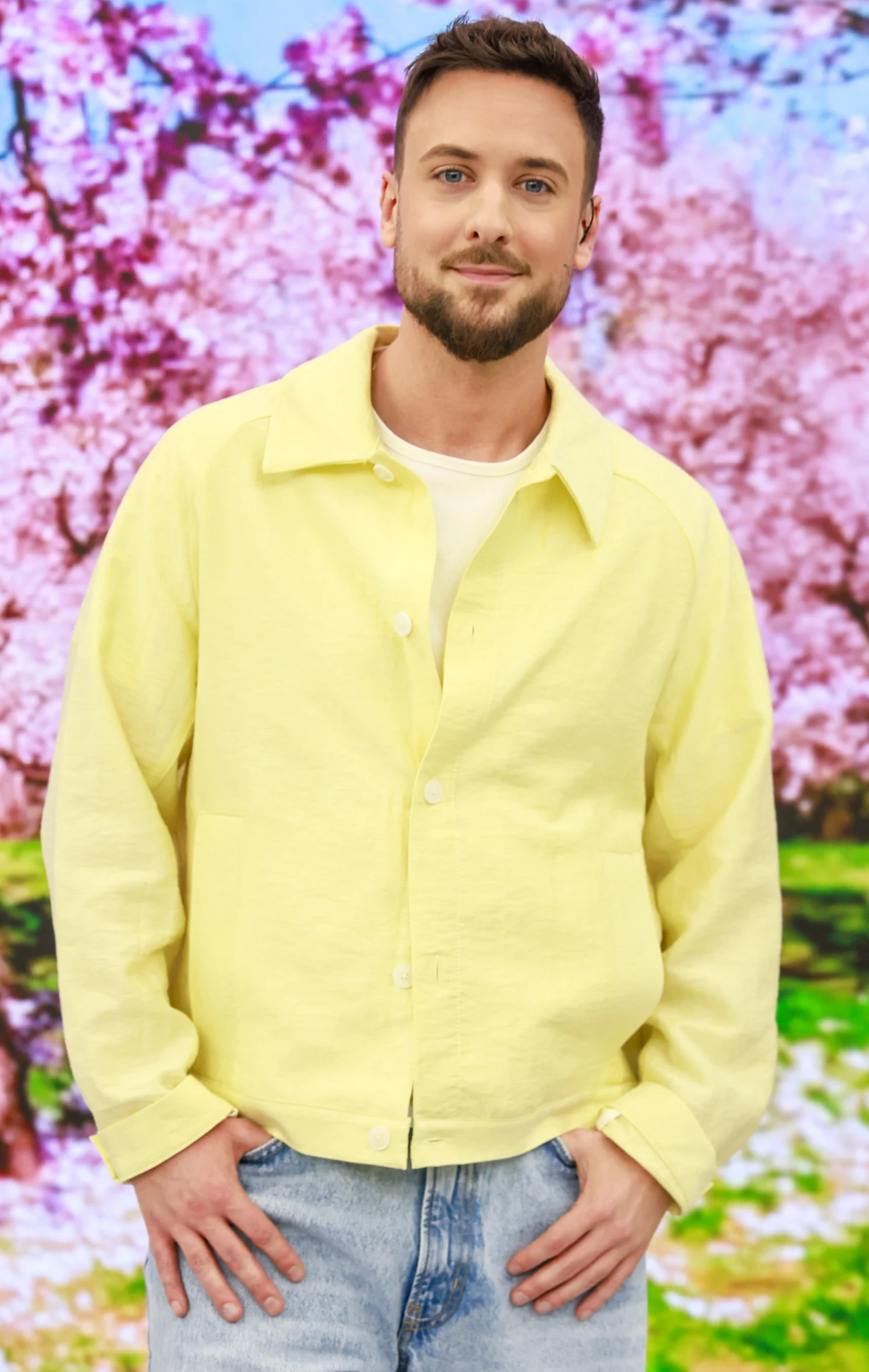 Aleksander Sikora w żółtej koszuli młody mężczyzna z krótką brodą ubrany w jasnożółtą koszulę i jasne dżinsy, stojący na tle kwitnących, różowych drzew w wiosennym parku
