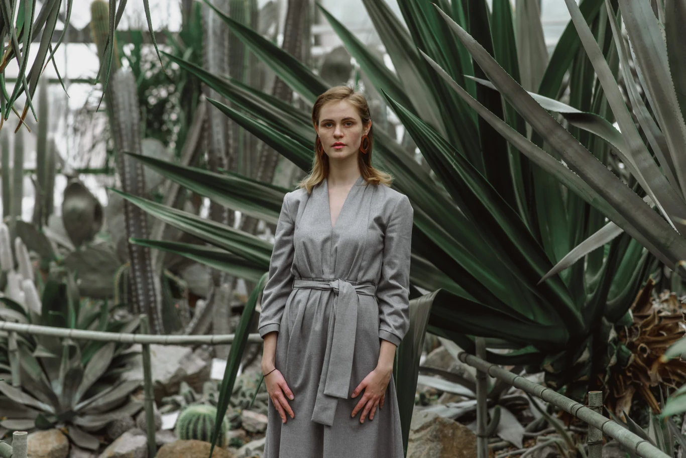 kobieta w szarej sukience stoi pośród dużych zielonych kaktusów i sukulentów w szklarni