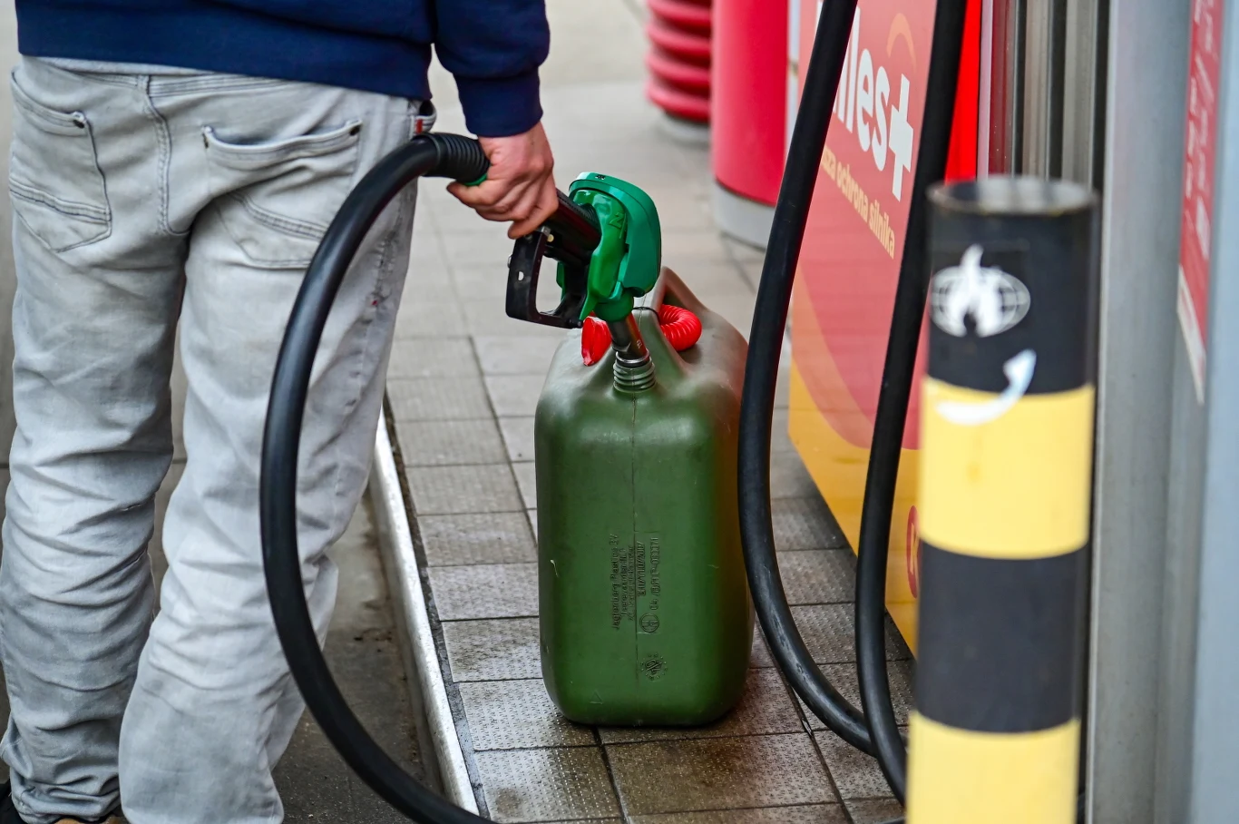 Mężczyzna w jeansach i niebieskiej bluzie tankuje zielony kanister przy dystrybutorze paliw na stacji benzynowej.