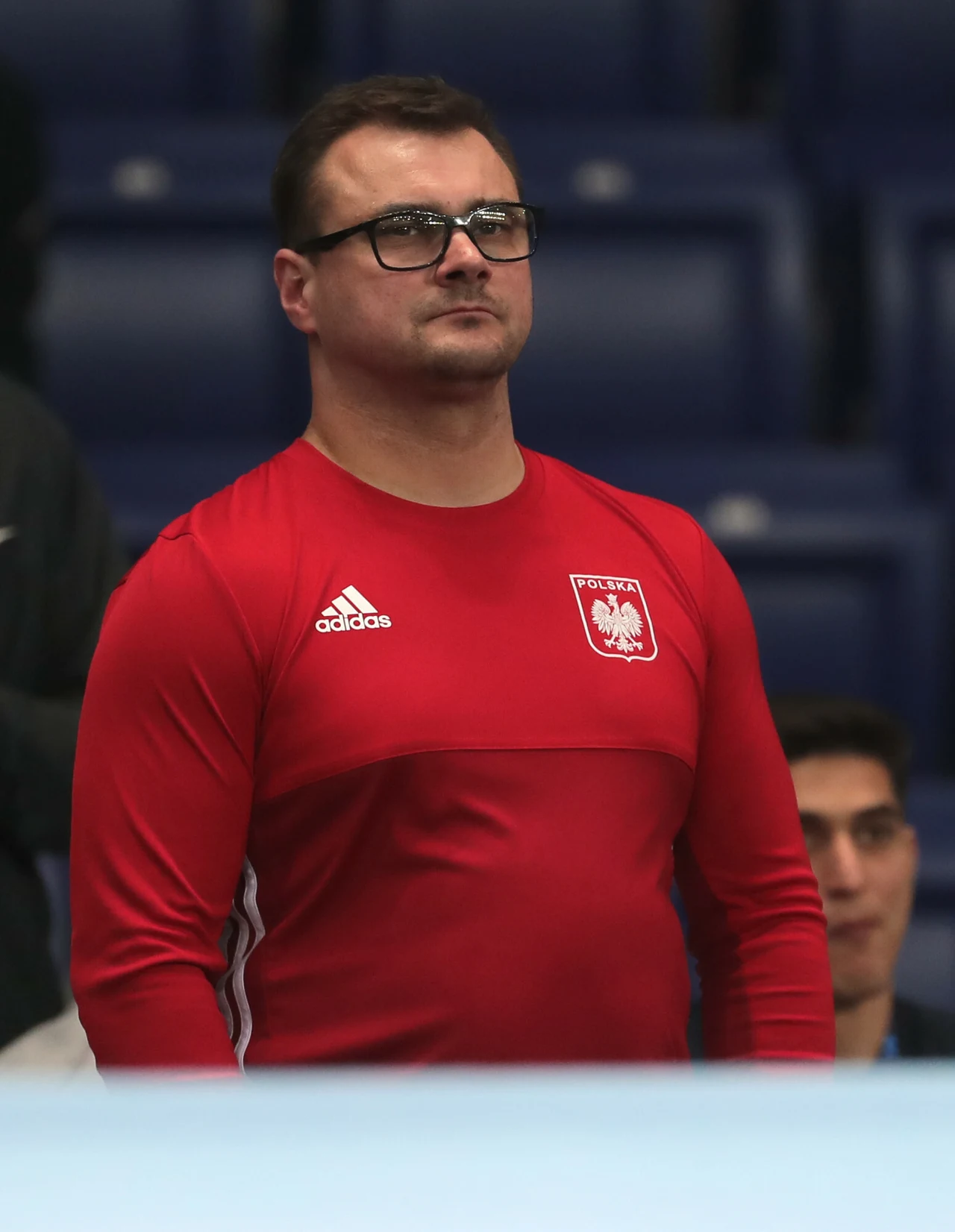 mężczyzna w okularach ubrany w czerwony sportowy dres z emblematem Polskiego Związku Piłki Nożnej i logo marki Adidas stoi na tle niebieskich trybun