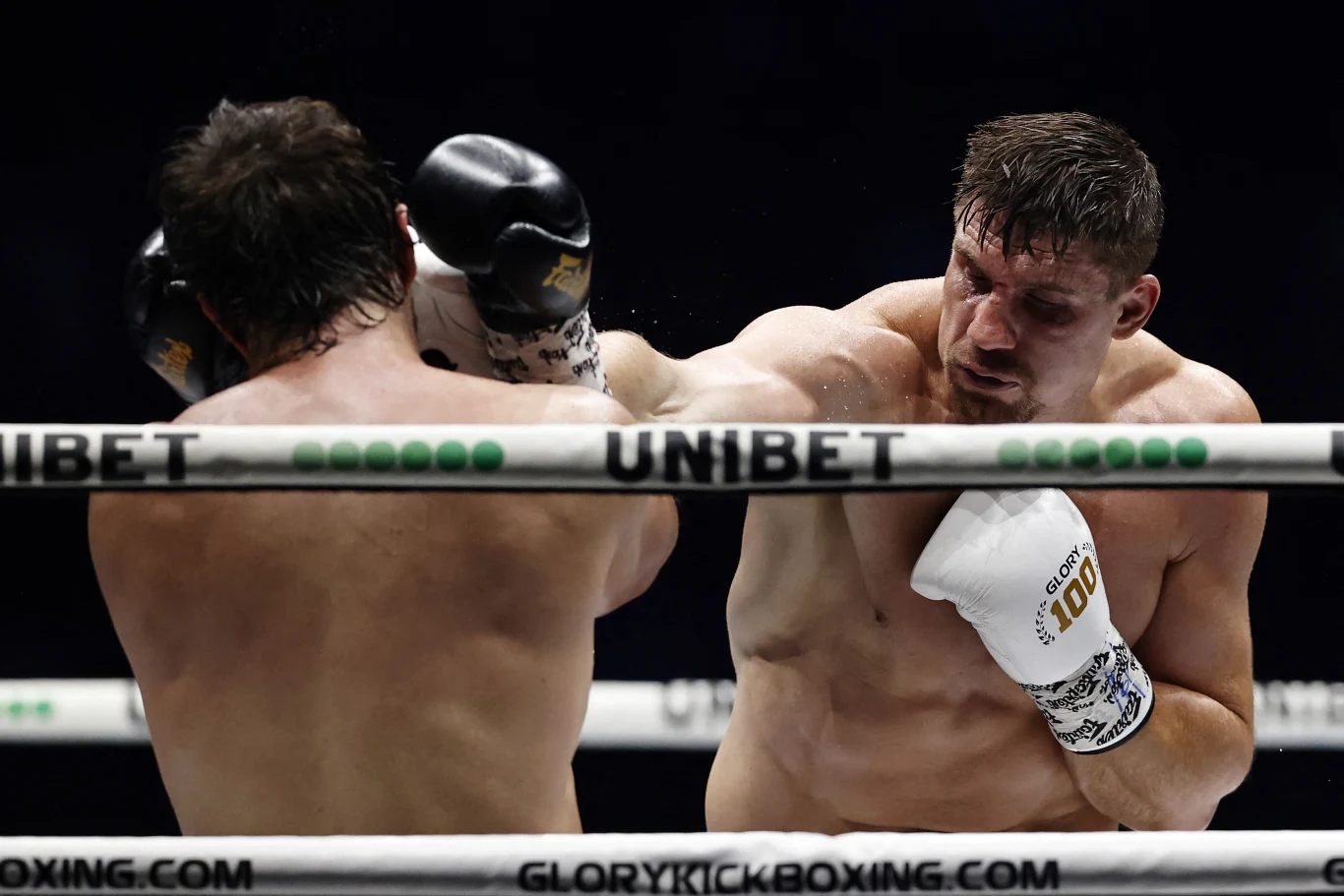 Dwóch bokserów w ringu podczas intensywnej wymiany ciosów, jeden z nich trafia rywala mocnym uderzeniem w twarz, w tle wyraźne liny ringu oraz logo sponsora Unibet.