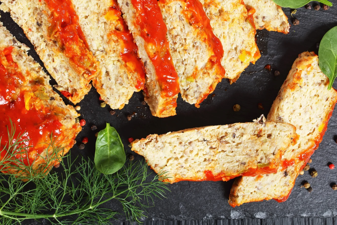 Pokrojony klops mięsny z sosem pomidorowym, udekorowany świeżym koperkiem, liśćmi szpinaku i kolorowym pieprzem, ułożony na czarnym talerzu.