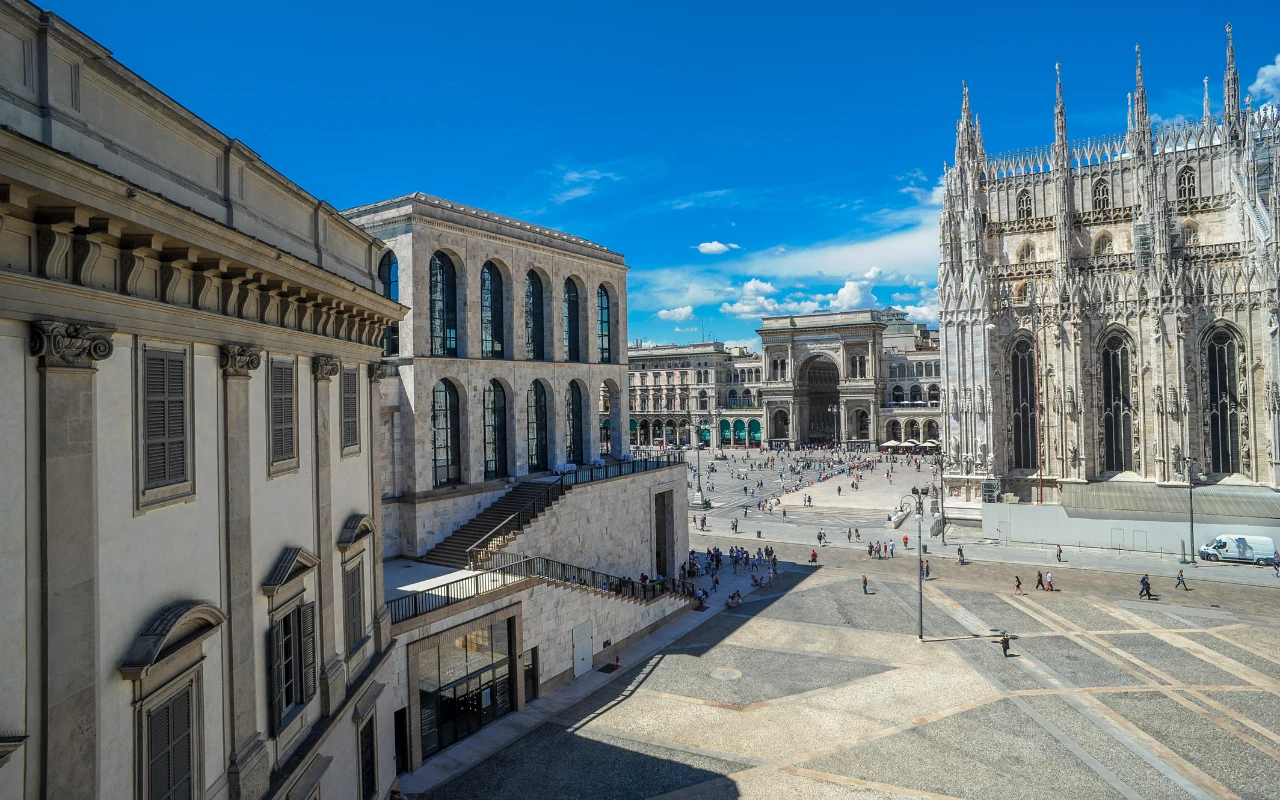 Główne elementy to monumentalna katedra z bogato zdobioną fasadą po prawej stronie oraz otwarty plac z niewielką liczbą spacerujących osób; w tle widoczny jest arkadowy budynek z przeszklonym przejściem i historyczne kamienice, wszystko otoczone jasnym...