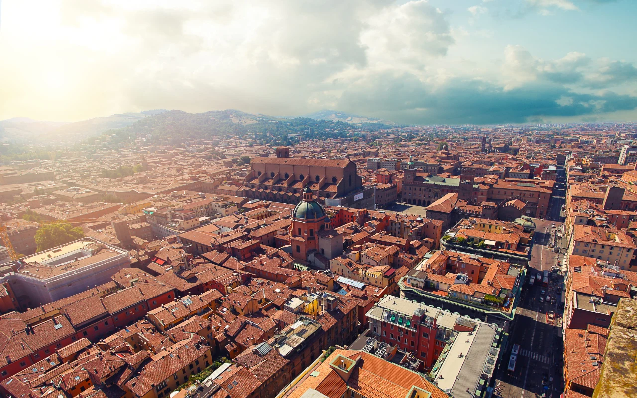 panorama miasta z charakterystycznymi czerwonymi dachami, w centrum widoczna duża katedra z kopułą, otoczona gęstą zabudową oraz prostokątnym placem, w tle rozciągają się wzgórza i chmury na niebie