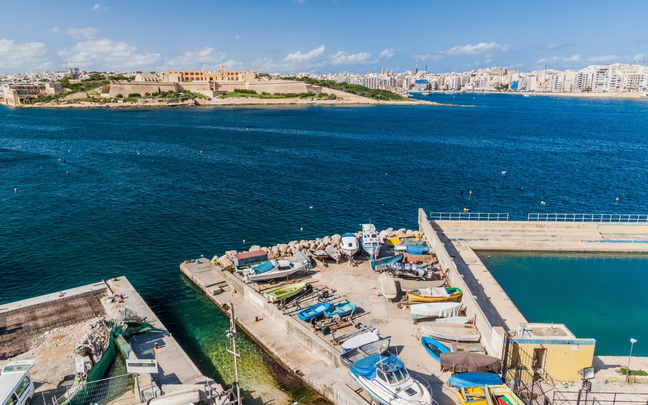 Niewielka stocznia z zacumowanymi łodziami i sprzętem rybackim położona przy błękitnej zatoczce, w tle szeroka panorama miasta oraz zabudowania na wzgórzu po drugiej stronie wody.