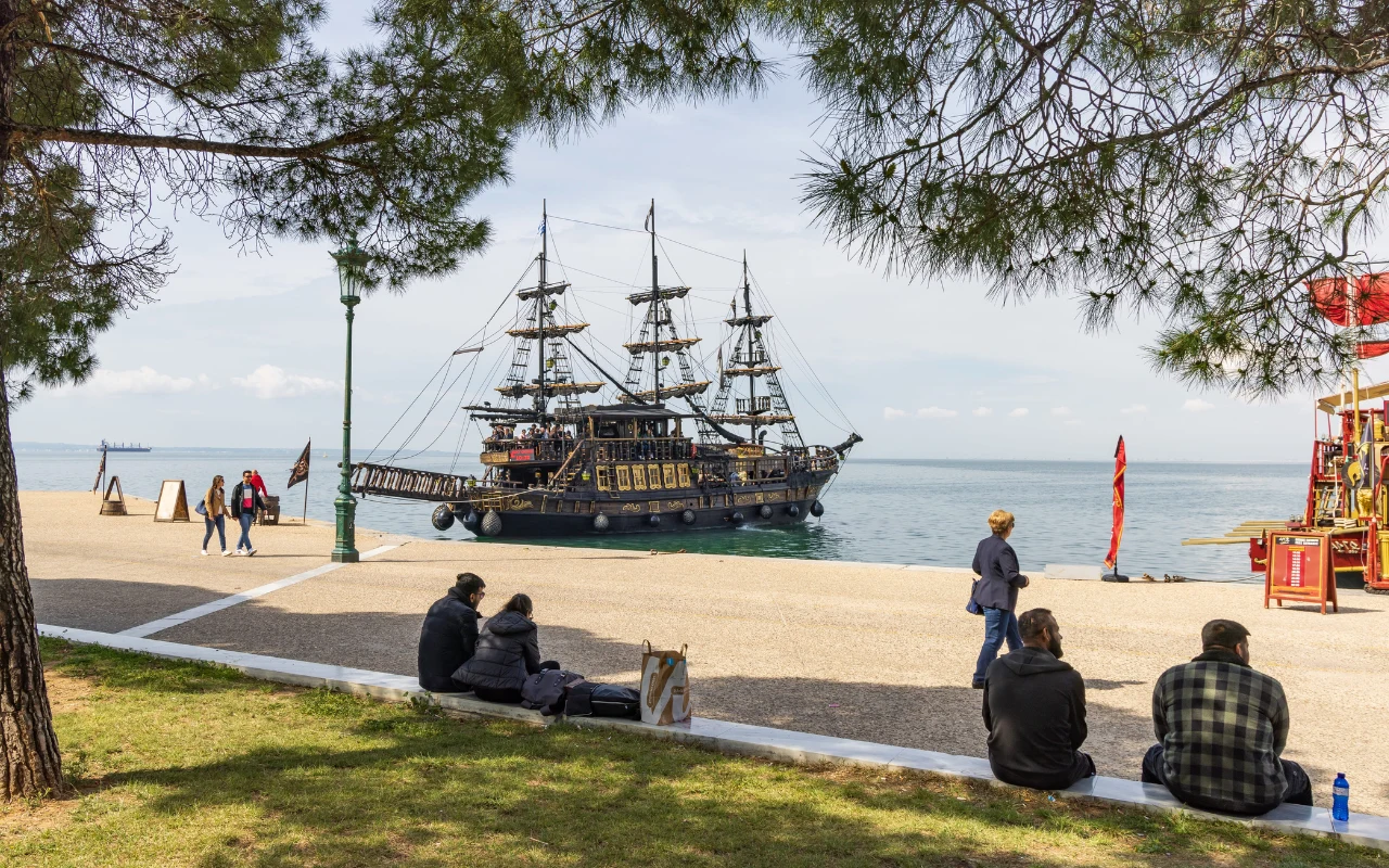 duży, czarny żaglowiec w stylu pirackim cumuje blisko brzegu, na pierwszym planie kilka osób siedzi na trawie i chodniku, patrząc w stronę statku, w tle spokojne morze i jasne niebo
