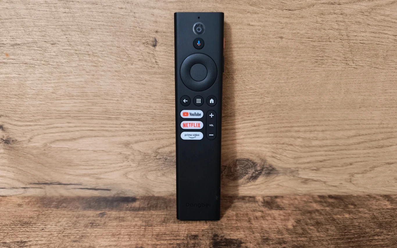 Czarny pilot z dużym okrągłym przyciskiem nawigacyjnym, przyciskami do sterowania głośnością oraz dedykowanymi przyciskami szybkiego dostępu do YouTube i Netflix na tle drewnianej powierzchni.