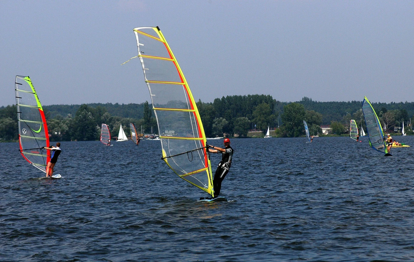Kilku windsurferów w piankach pływających na desce z żaglem po jeziorze, otoczonym przez drzewa i niewielką liczbę budynków na horyzoncie.