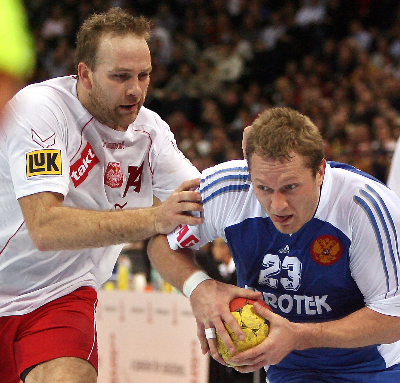 dwóch zawodników podczas meczu piłki ręcznej, jeden w białym stroju z orłem na piersi próbuje powstrzymać przeciwnika w niebieskim stroju z piłką w rękach