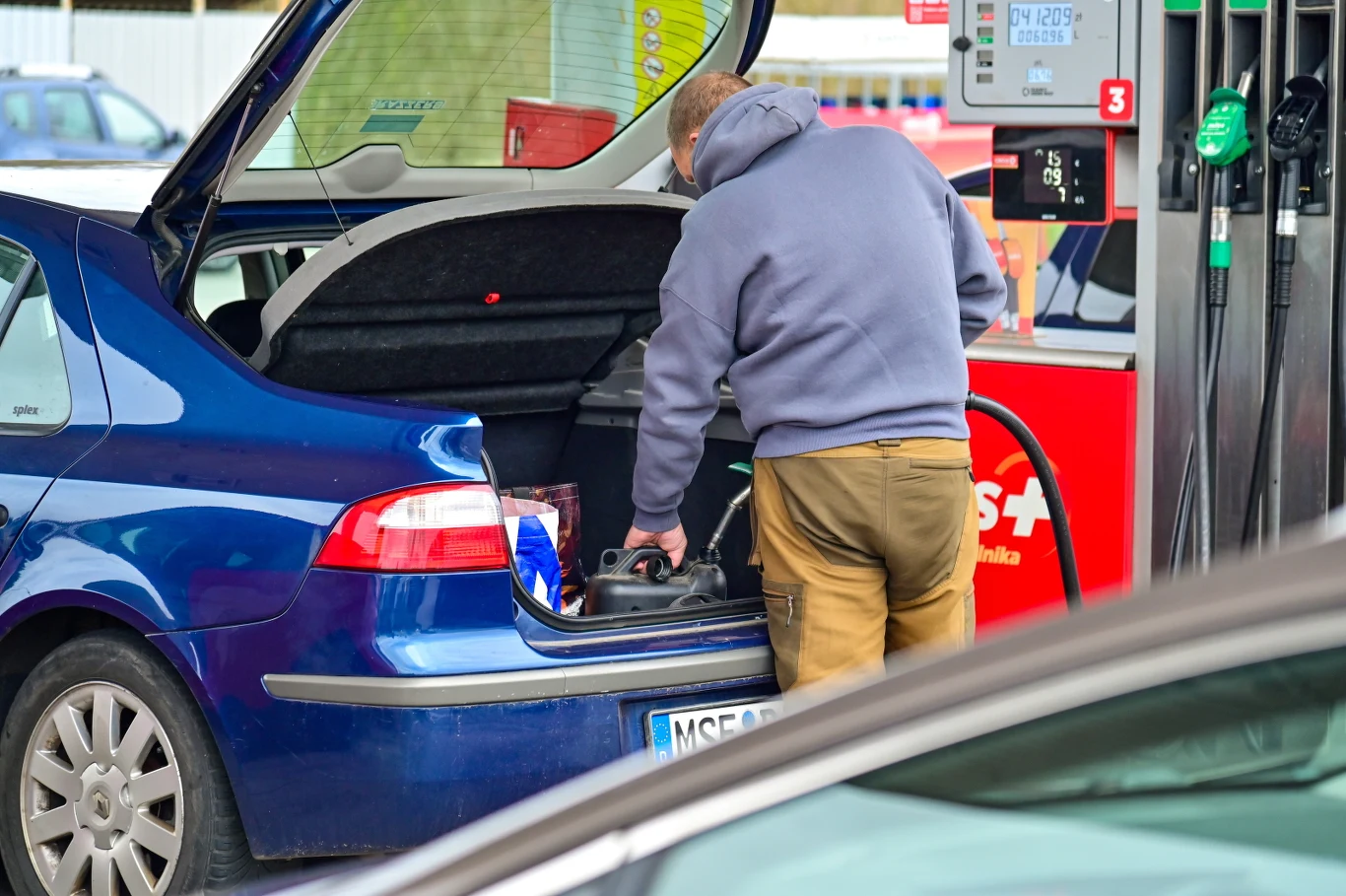 mężczyzna w sportowej bluzie pochylony nad bagażnikiem niebieskiego samochodu na stacji paliw, wkładający kanister lub narzędzie do środka