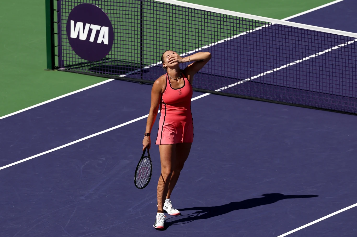 tenisistka w różowym stroju sportowym stoi na korcie, trzyma rakietę w jednej ręce, drugą zakrywa twarz w geście rozczarowania lub frustracji; w tle siatka oraz logo WTA