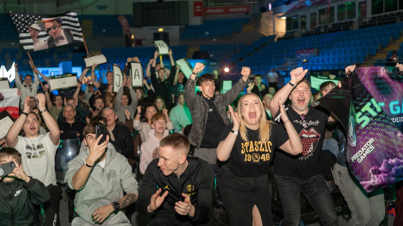 Grupa rozentuzjazmowanych osób kibicujących na wydarzeniu e-sportowym, trzymających transparenty i flagi, z widocznymi emocjami i podnoszonymi rękami na tle trybun hali sportowej.