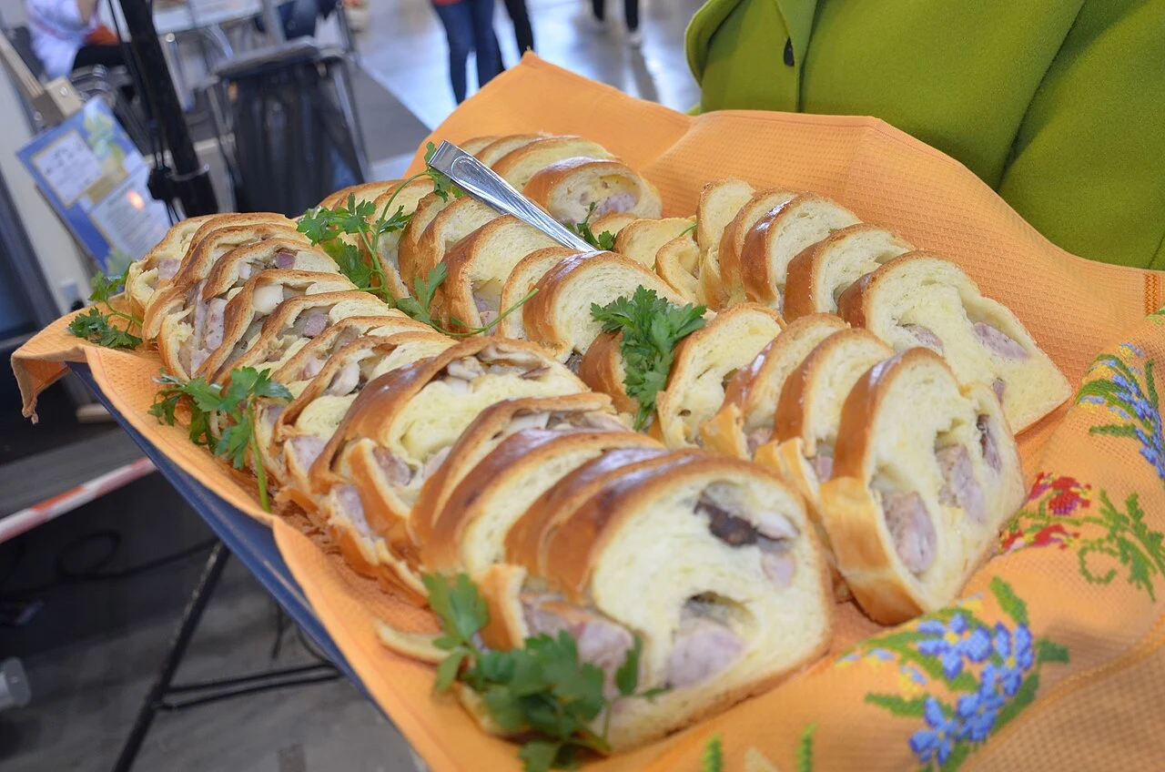 Pokrojone w plastry drożdżowe ciasto z nadzieniem mięsnym, podane na pomarańczowej serwetce ozdobionej haftem, udekorowane świeżą natką pietruszki, z włożonym srebrnym nożem.