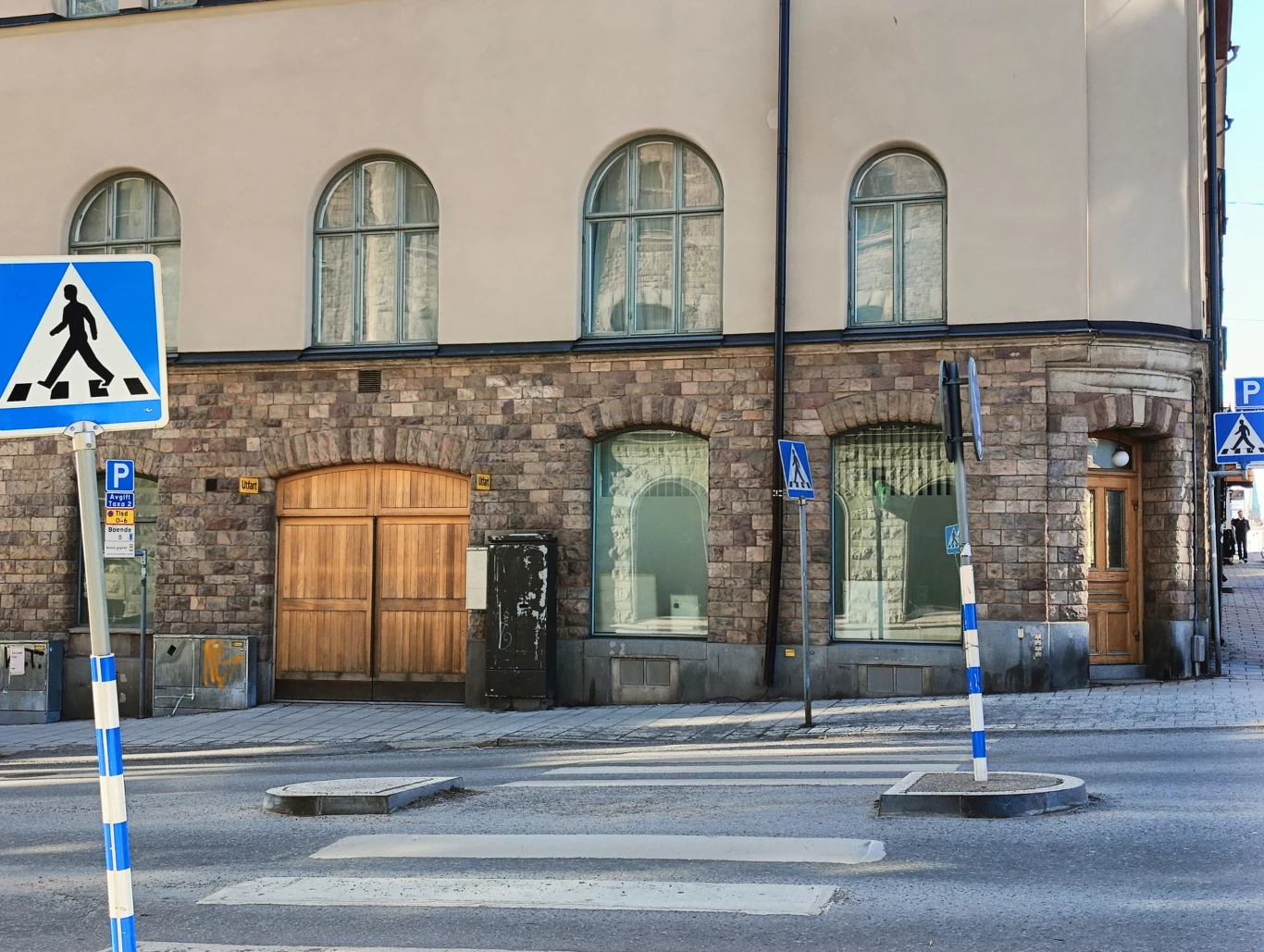 Skrzyżowanie z przejściem dla pieszych prowadzącym do kamienicy z dużymi oknami i drzwiami z jasnego drewna, otoczonej znakami drogowymi i niebiesko-białymi słupkami przy krawężniku.
