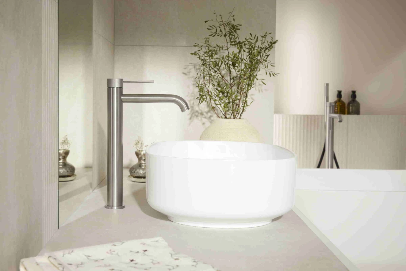 nowoczesna umywalka nablatowa w kształcie misy, srebrna bateria, jasny blat oraz duży ozdobny wazon z zieloną rośliną