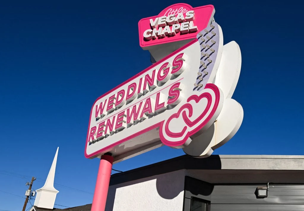 kolorowy neonowy szyld z napisem „Little Vegas Chapel” oraz „Weddings Renewals” z motywem dwóch splecionych serc, umieszczony na tle błękitnego nieba nad budynkiem; po lewej stronie fragment iglicy innego budynku
