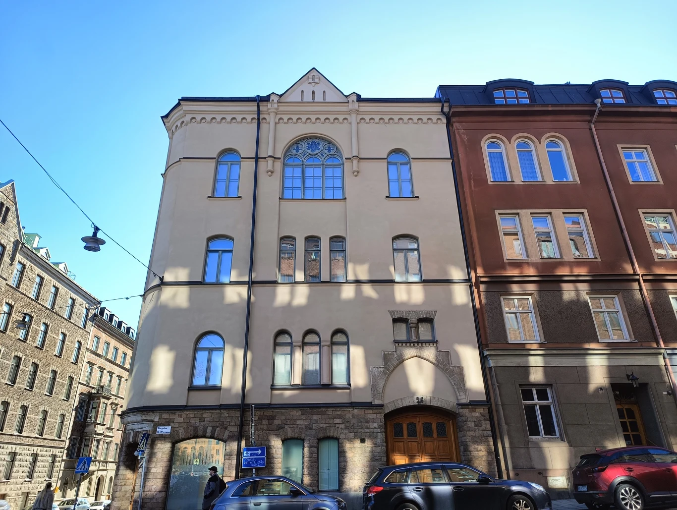 Jasna kamienica z dużym, półokrągłym oknem na piętrze, otoczona wyższymi budynkami z cegły, na ulicy zaparkowane samochody, a na chodniku widoczny przechodzień.