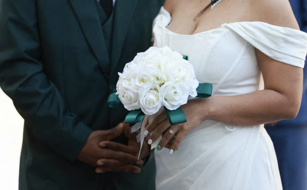 Para młoda w eleganckich strojach trzymająca bukiet białych róż z zieloną wstążką, podkreślających uroczysty charakter chwili.