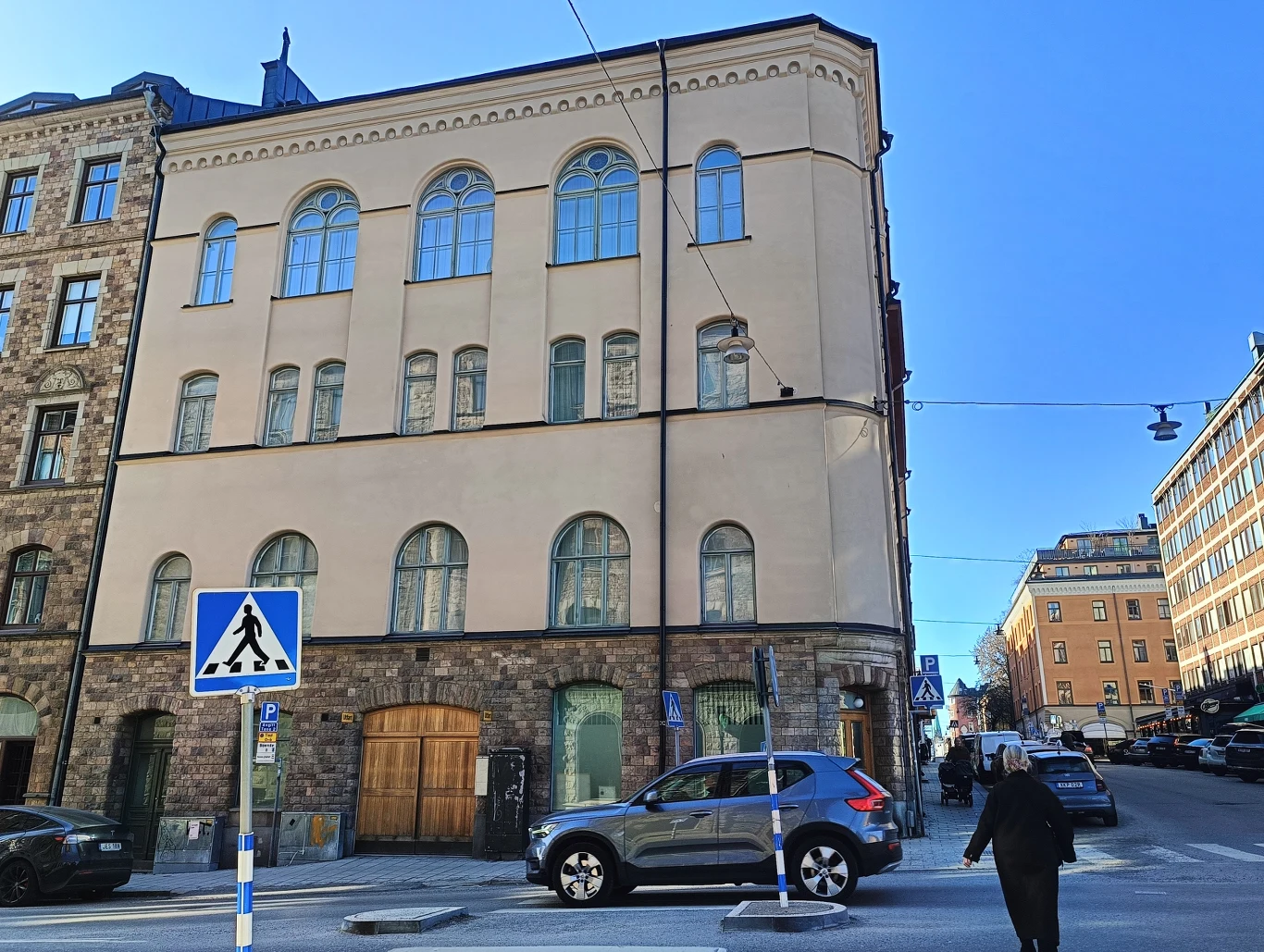 zabytkowa kamienica z jasną elewacją i dużymi, łukowatymi oknami, u jej podnóża zaparkowane samochody, na pierwszym planie przejście dla pieszych, kobieta przechodząca przez ulicę, niebo bezchmurne i jasne