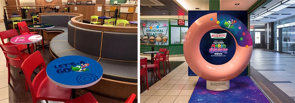 Kolorowe stoliki i krzesła w kawiarni z motywem gry lub kreskówki, a obok dekoracyjna instalacja w kształcie dużego pączka z grafiką promującą akcję Krispy Kreme i kosmiczne motywy.