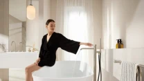 kobieta w czarnym szlafroku siedząca na krawędzi wolnostojącej wanny, wypełniająca ją wodą w jasnej, nowoczesnej łazience z minimalistycznym wystrojem, obok których znajduje się stylowy stojak z ręcznikiem i kosmetykami