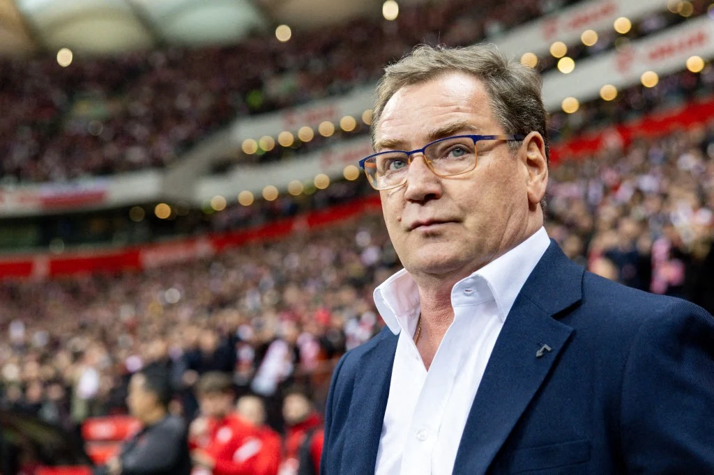 mężczyzna w okularach i eleganckim garniturze stoi na tle wypełnionego kibicami stadionu, skupiony wyraz twarzy sugeruje zaangażowanie w wydarzenie sportowe