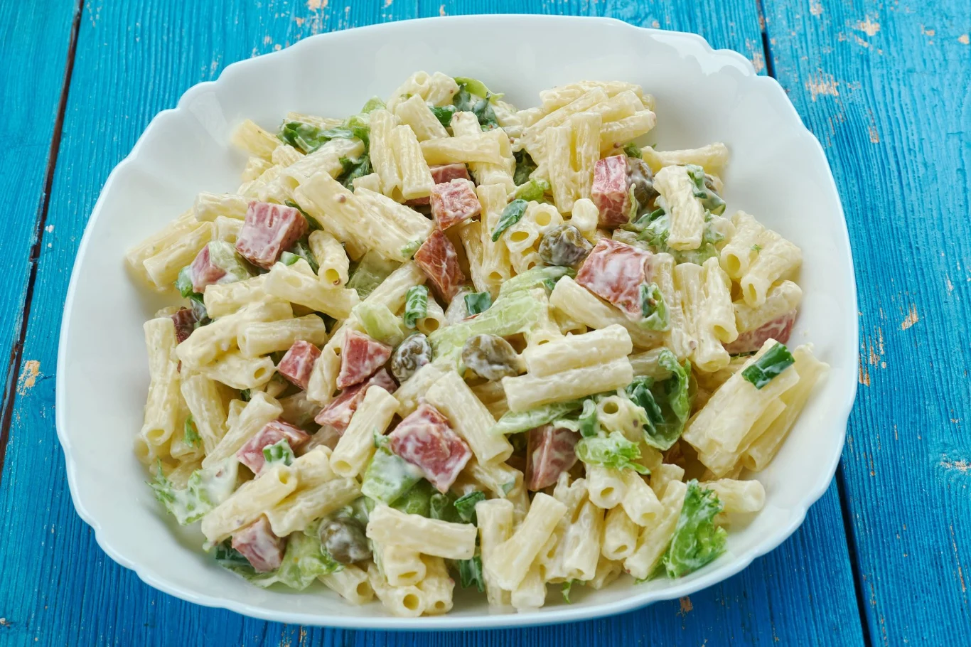 Pyszna sałatka makaronowa. Syci na długi czas Sałatka makaronowa z rurkami, szynką, sałatą i majonezem w białej misce na niebieskim stole