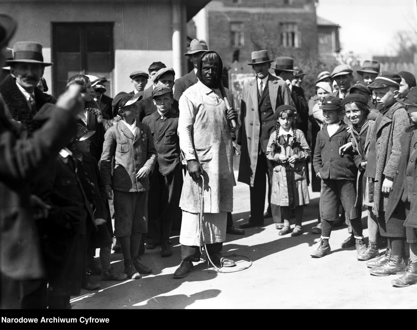 1935 rok: Siuda Baba to regionalny zwyczaj wielkanocny w Wieliczce Czarnoskóry mężczyzna w długim jasnym fartuchu i chuście na głowie stoi z biczem lub pejczem w ręku pośrodku kręgu osób w kapeluszach i garniturach oraz dzieci, które z zainteresowaniem obserwują go z różnych stron.