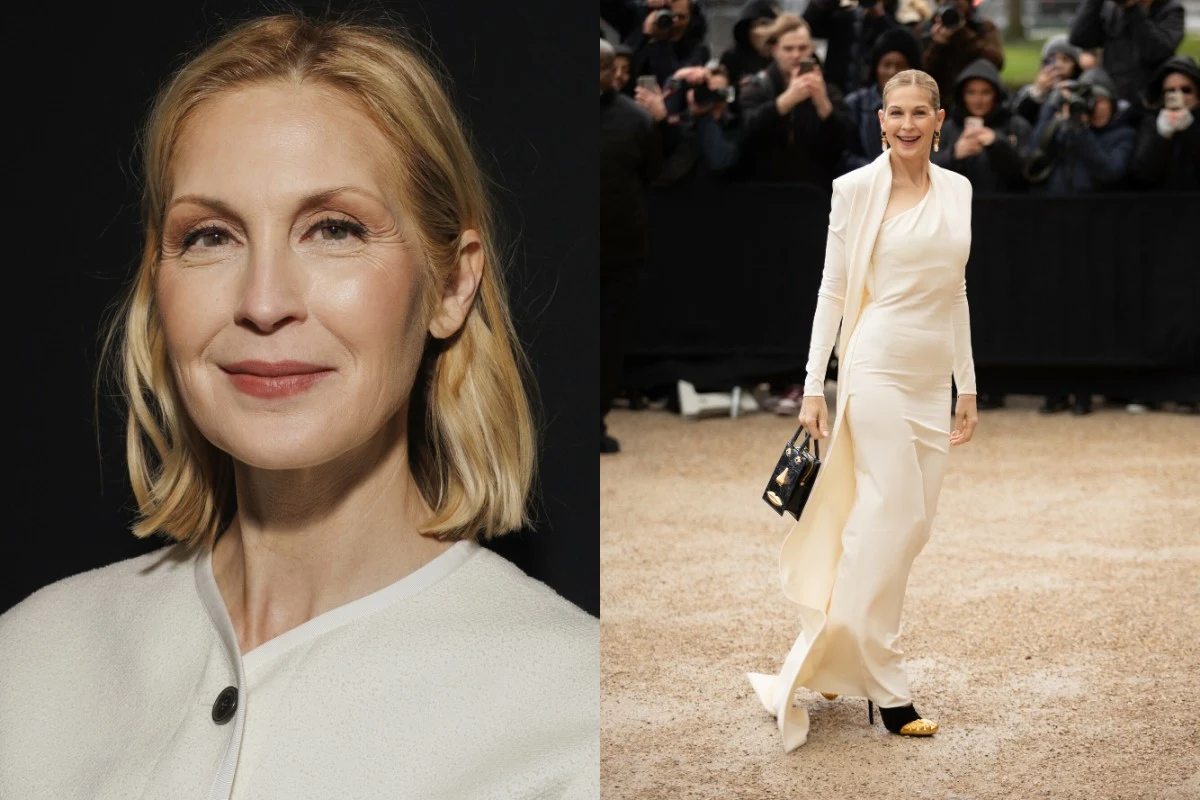 kobieta o jasnych, blond włosach w eleganckiej, białej stylizacji, z uśmiechem na twarzy, pozuje na tle tłumu fotografów, trzymając czarną torebkę i stojąc na jasnym podłożu