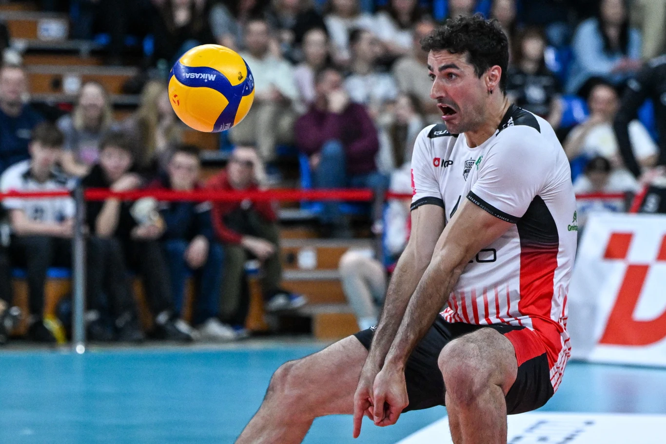 siatkarz w białej koszulce i czerwonych spodenkach odbija piłkę do siatkówki, koncentrując się na precyzyjnym przyjęciu, a w tle widoczna jest grupa kibiców na trybunach