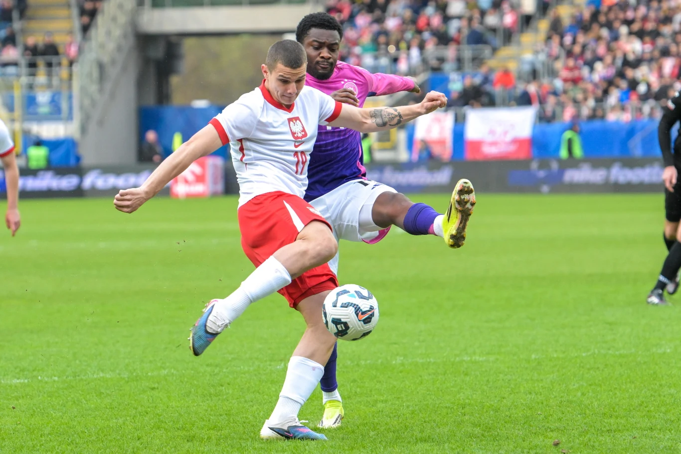 dwóch piłkarzy walczy o piłkę na murawie podczas meczu, zawodnik w białoczerwonym stroju wybija piłkę sprzed przeciwnika w fioletowym stroju, na trybunach liczna publiczność