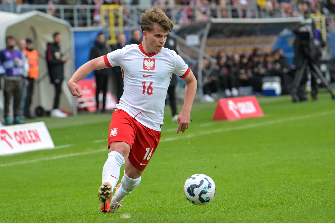 młody piłkarz w białej koszulce z numerem 16 i czerwonych spodenkach biegnie przy linii bocznej boiska, koncentrując się na piłce nożnej podczas meczu; w tle widoczna publiczność na trybunach