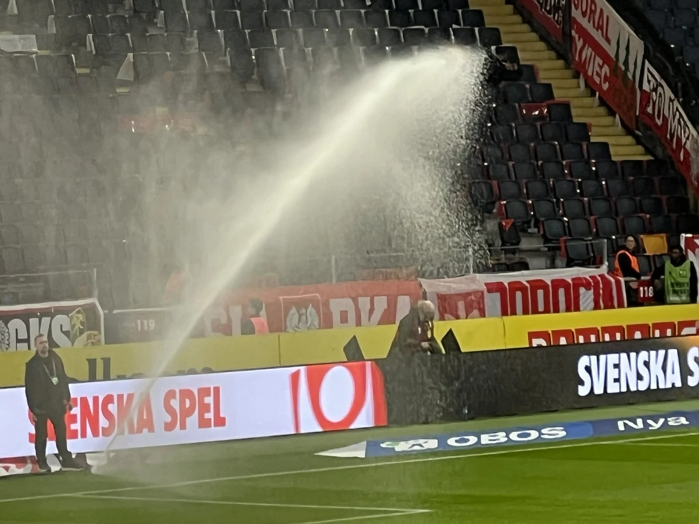 silny strumień wody z węża podlewającego murawę na stadionie kieruje się bezpośrednio na stojącą przy bandzie osobę, która jest obficie zraszana, w tle puste trybuny i banery reklamowe