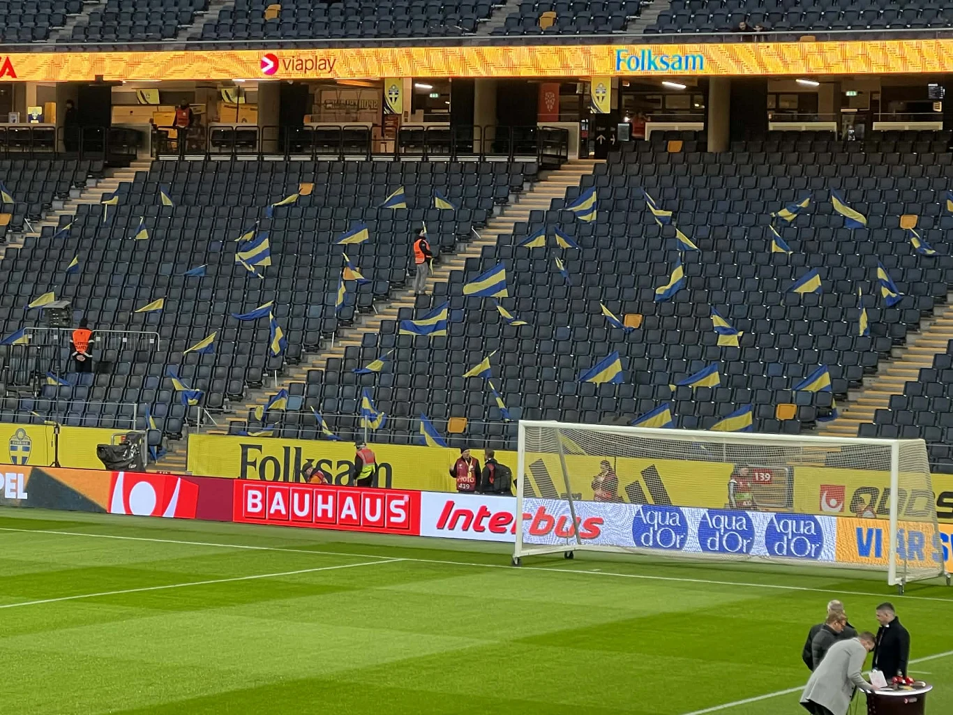 trybuna stadionu z żółto-niebieskimi flagami rozłożonymi na krzesełkach, bramką piłkarską na pierwszym planie oraz kilkoma osobami pracującymi przy murawie