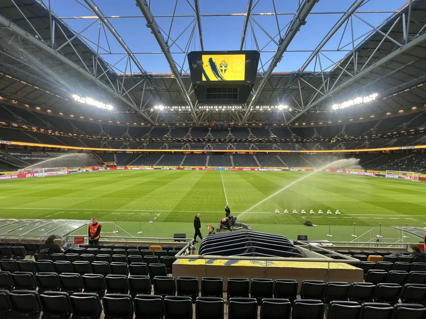 nowoczesny stadion piłkarski z zamkniętym dachem, podlewaną murawą oraz dużym ekranem zawieszonym nad boiskiem, na trybunach pojedyncze osoby przygotowujące się do wydarzenia