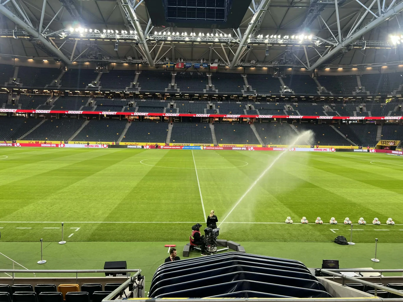 zielona murawa stadionu piłkarskiego oświetlona sztucznym światłem, na której działa zraszacz podlewający trawę; na pierwszym planie znajduje się kamera telewizyjna obsługiwana przez operatora, a trybuny w tle są niemal puste
