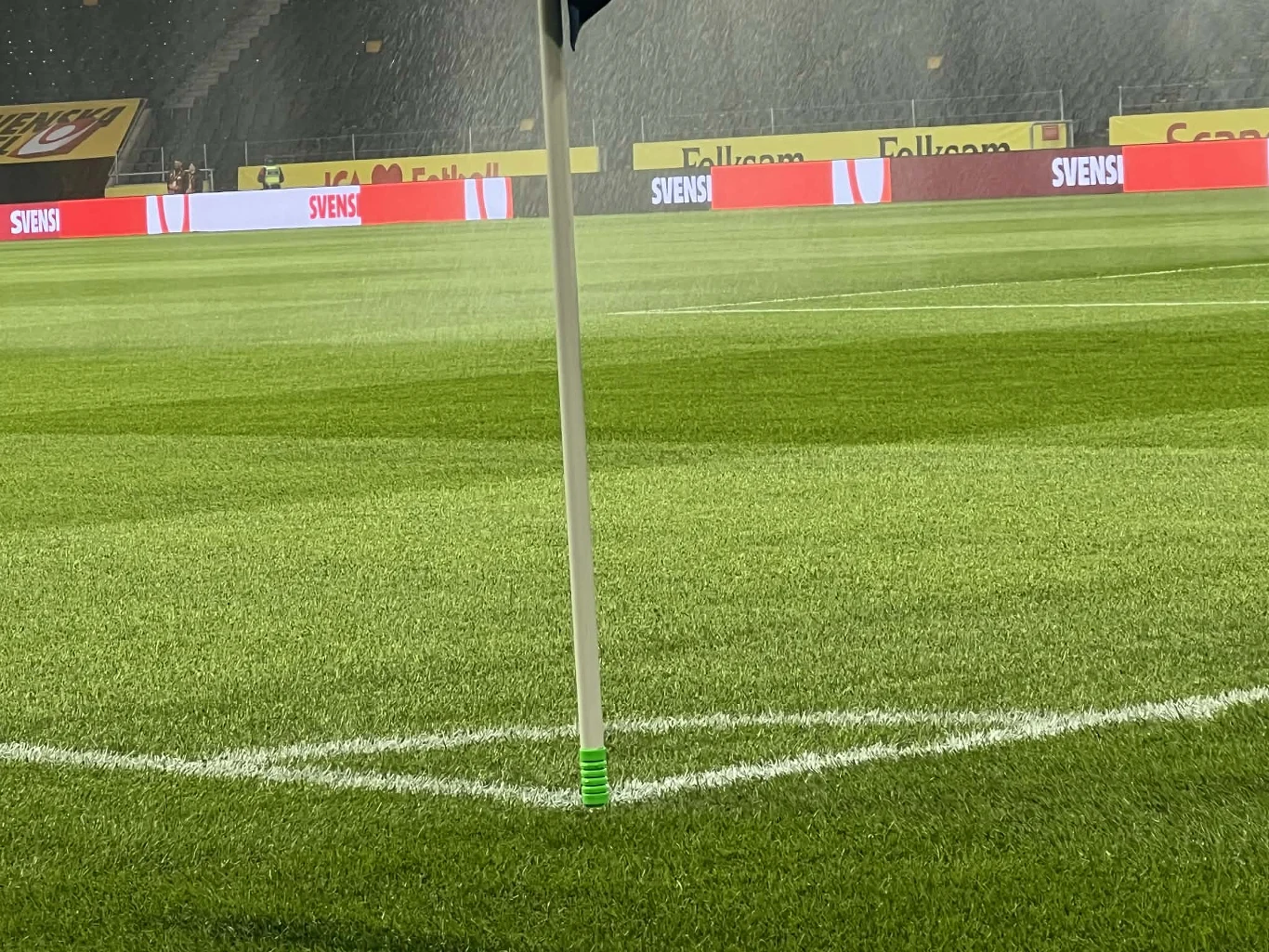 chorągiewka narożna ustawiona w rogu boiska piłkarskiego na tle równo przystrzyżonej, zielonej murawy z wyraźnie widocznymi liniami pola gry, w oddali jasne reklamy na bandach stadionowych