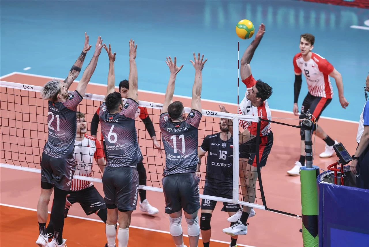 zawodnik w czerwono-białym stroju wykonuje atak nad siatką, podczas gdy trzech graczy w ciemnych koszulkach skacze do bloku; piłka znajduje się nad rękami atakującego, a na boisku widoczni są także inni zawodnicy przygotowani do akcji