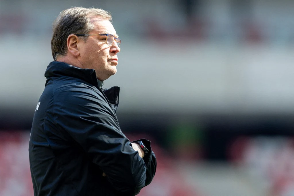 mężczyzna w średnim wieku w okularach i czarnej kurtce stoi bokiem z założonymi rękami, skupiony patrzy przed siebie