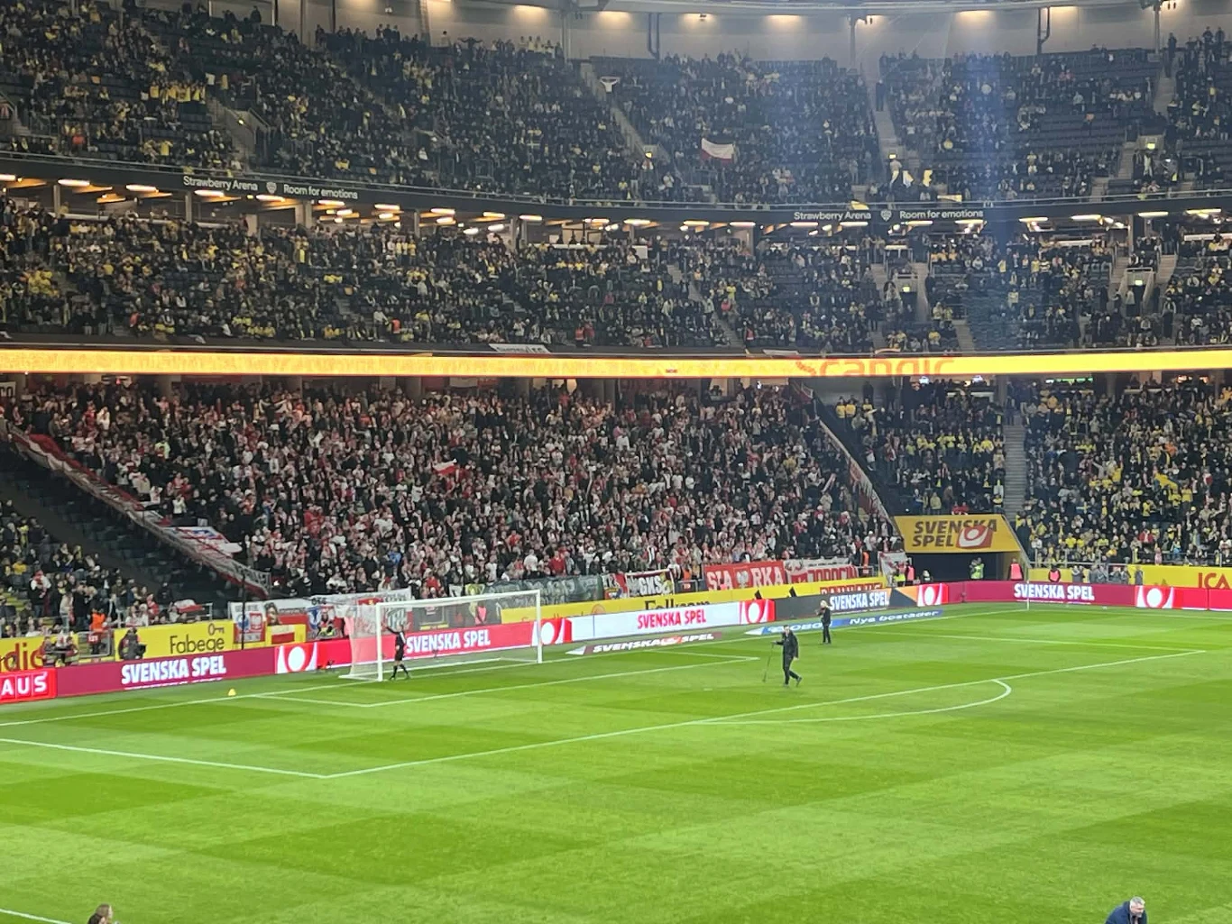 Tłum kibiców ubranych w żółte i czerwono-białe barwy zajmuje trybuny dużego stadionu, skupiając się za bramką, a centralnie na murawie znajduje się sędzia oraz fragment boiska przygotowany do rozegrania meczu piłki nożnej.