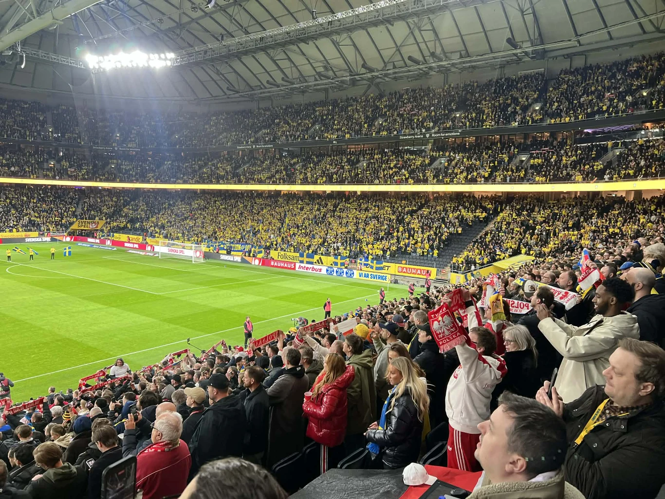 Tłum kibiców na stadionie piłkarskim, sektor kibiców w biało-czerwonych barwach aktywnie dopinguje swoją drużynę, podczas gdy reszta trybun dominuje w żółtych koszulkach, tworząc wyraźny kontrast między grupami fanów.