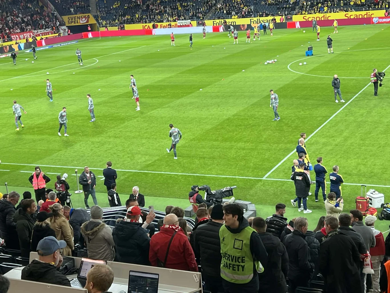 piłkarze wykonują rozgrzewkę na murawie stadionu, wokół boiska poruszają się sędziowie, pracownicy techniczni i ochrona, a na trybunach gromadzi się publiczność; w tle ławki rezerwowych i telewizyjne kamery