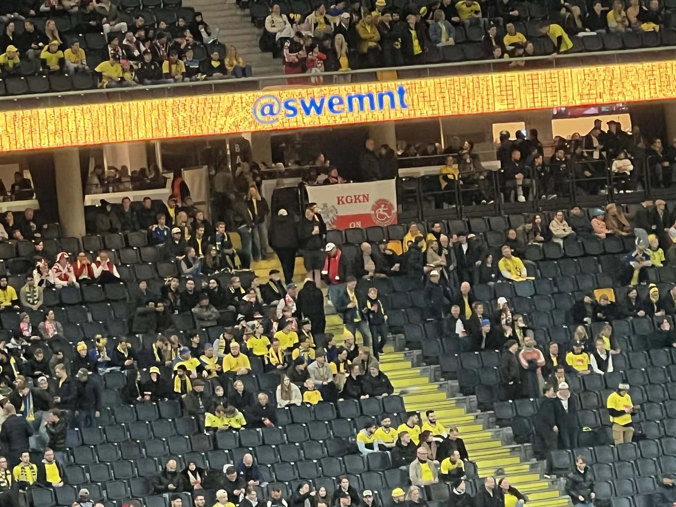 tłum kibiców na trybunach, dominują żółte barwy ubrań, w centralnej części widoczna polska flaga z napisem 'Kęty'; nad miejscami widnieje podświetlany napis '@swemnt'